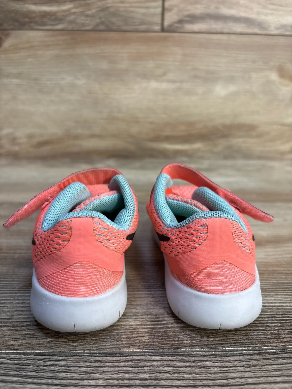 Rear view of kids coral pink mesh sneakers showcasing aqua collar lining and white rubber soles on wood plank floor