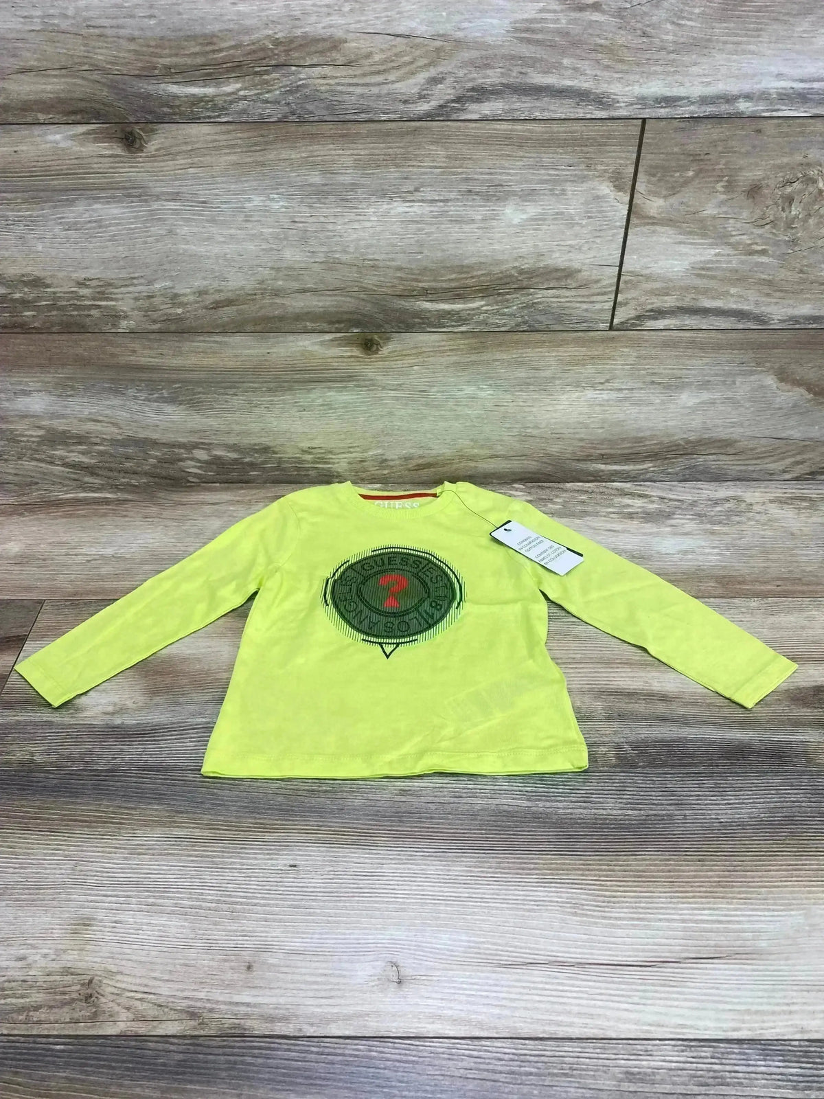 Front view of size 2T Guess long-sleeve toddler shirt showcasing round logo print on chest with wood plank background