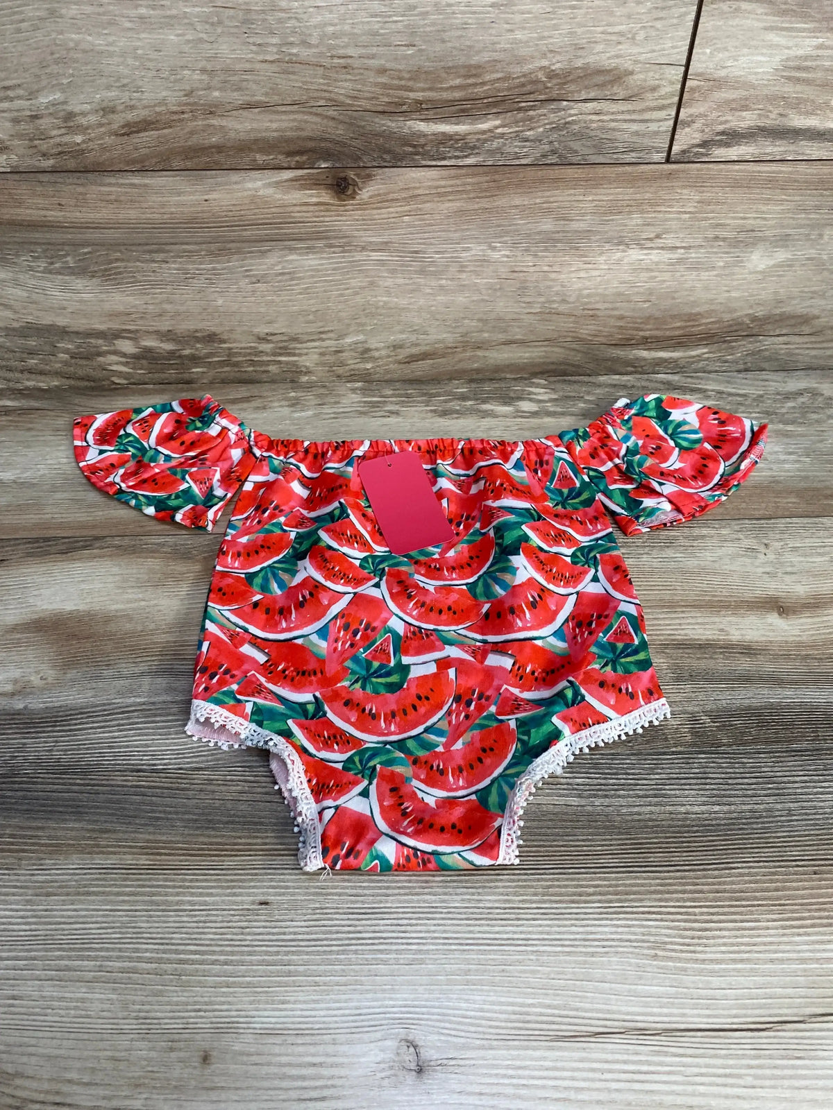 Front view of red watermelon-print romper showcasing an off-shoulder ruffled neckline and lace-trimmed leg openings on a wood surface