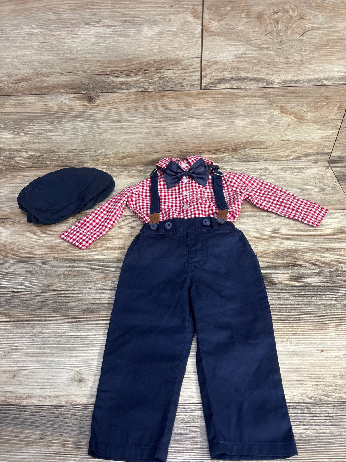 Children's outfit with red checkered shirt, navy blue pants with suspenders, bow tie, and matching navy cap on wooden floor