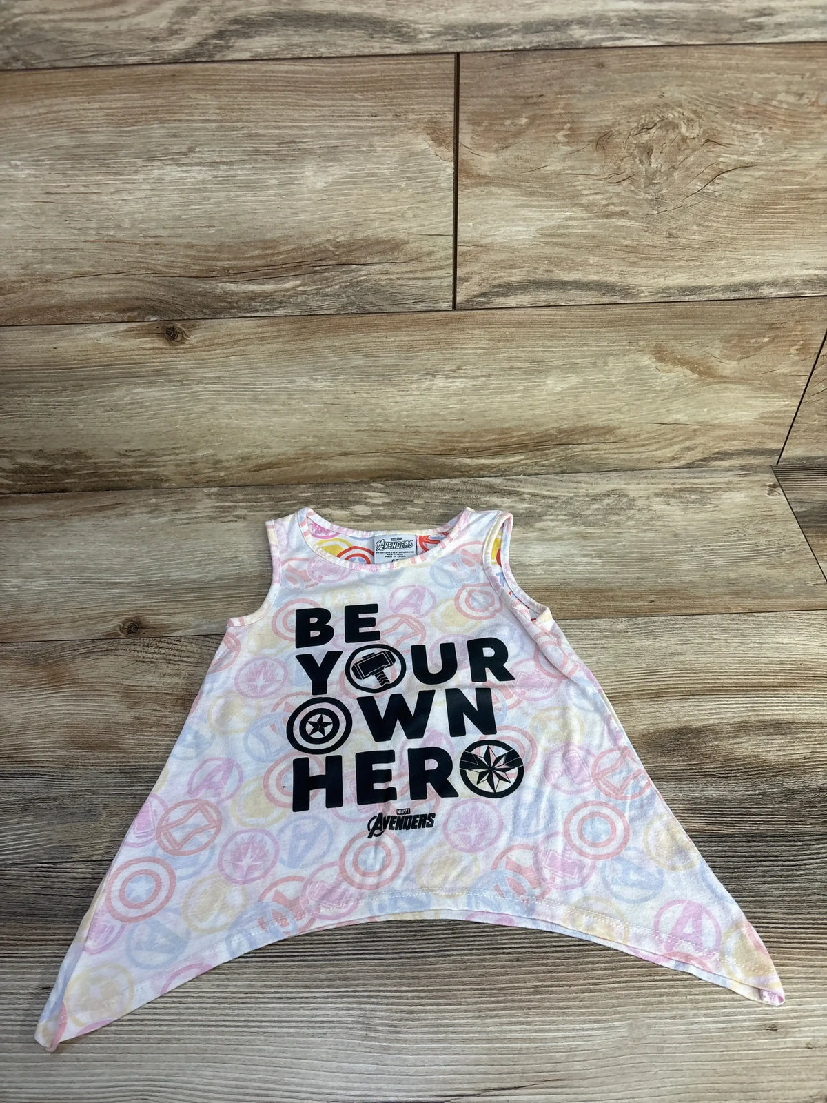 Flat lay of white Avengers flare tank top with black BE YOUR OWN HERO print and pastel circular icons on wood
