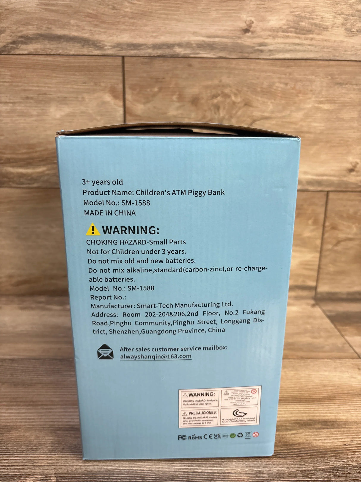 Rear view of children's ATM piggy bank box showing warning label and product information on a light blue box against a wooden floor background