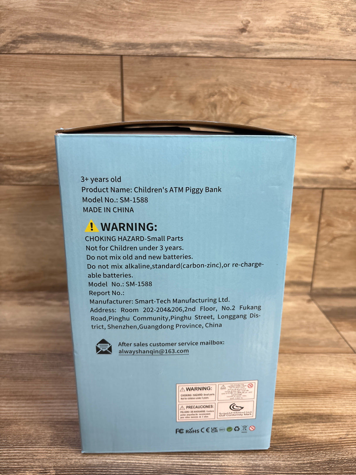 Rear of Fishboy Talking ATM Piggy Bank box showing WARNING: label and product info on light blue background over wooden floor