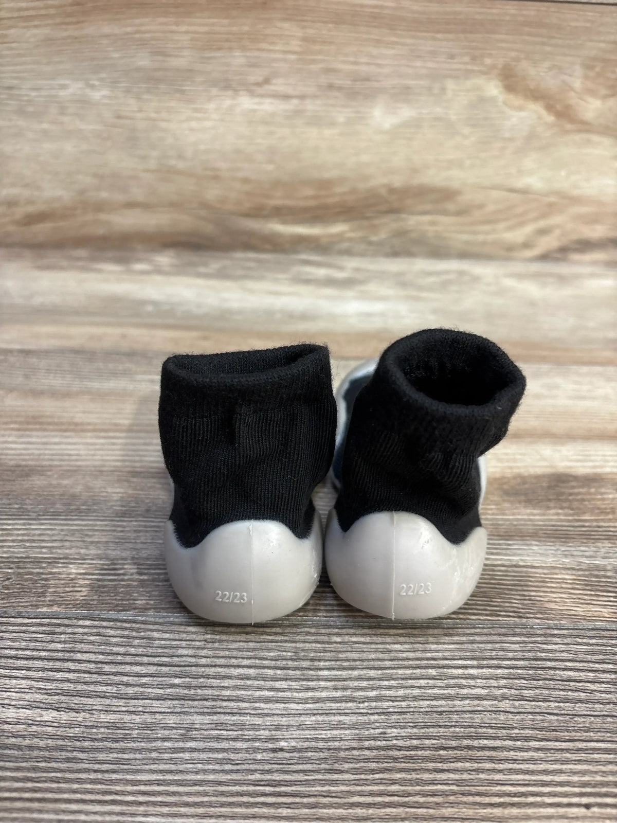 Rear view of black baby sock shoes showcasing white rubber soles marked 22 23 on a wood background