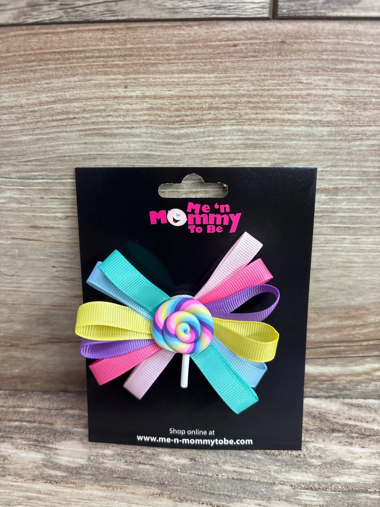 Front view of lollipop applique hair bow showcasing multicolor ribbon loops and rainbow lollipop center on a black display card with Me 'n Mommy To Be logo