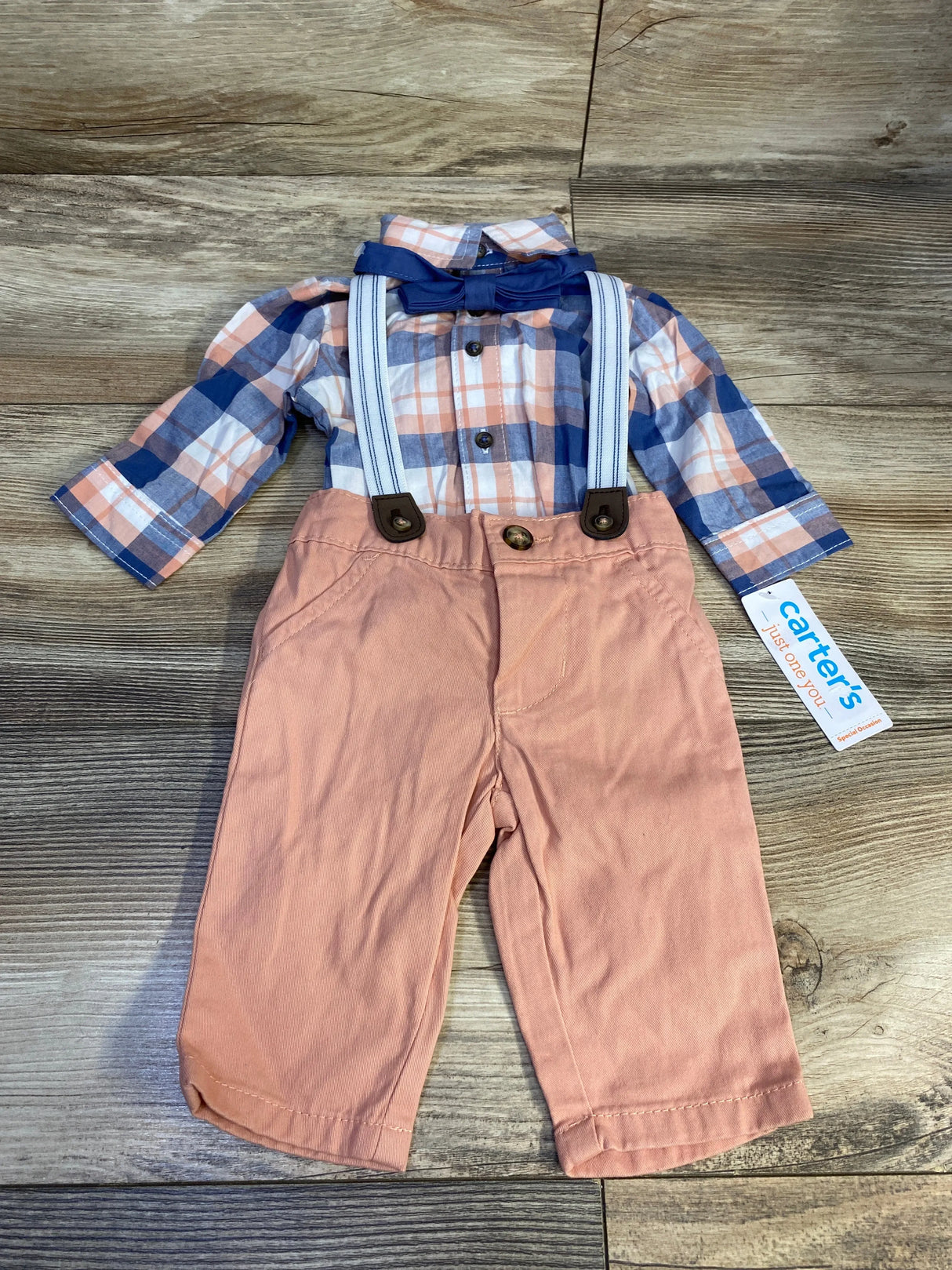 Flat lay of 4-piece baby plaid button-up shirt with navy bow tie and pink pants with white-striped suspenders on wood background, size 3M