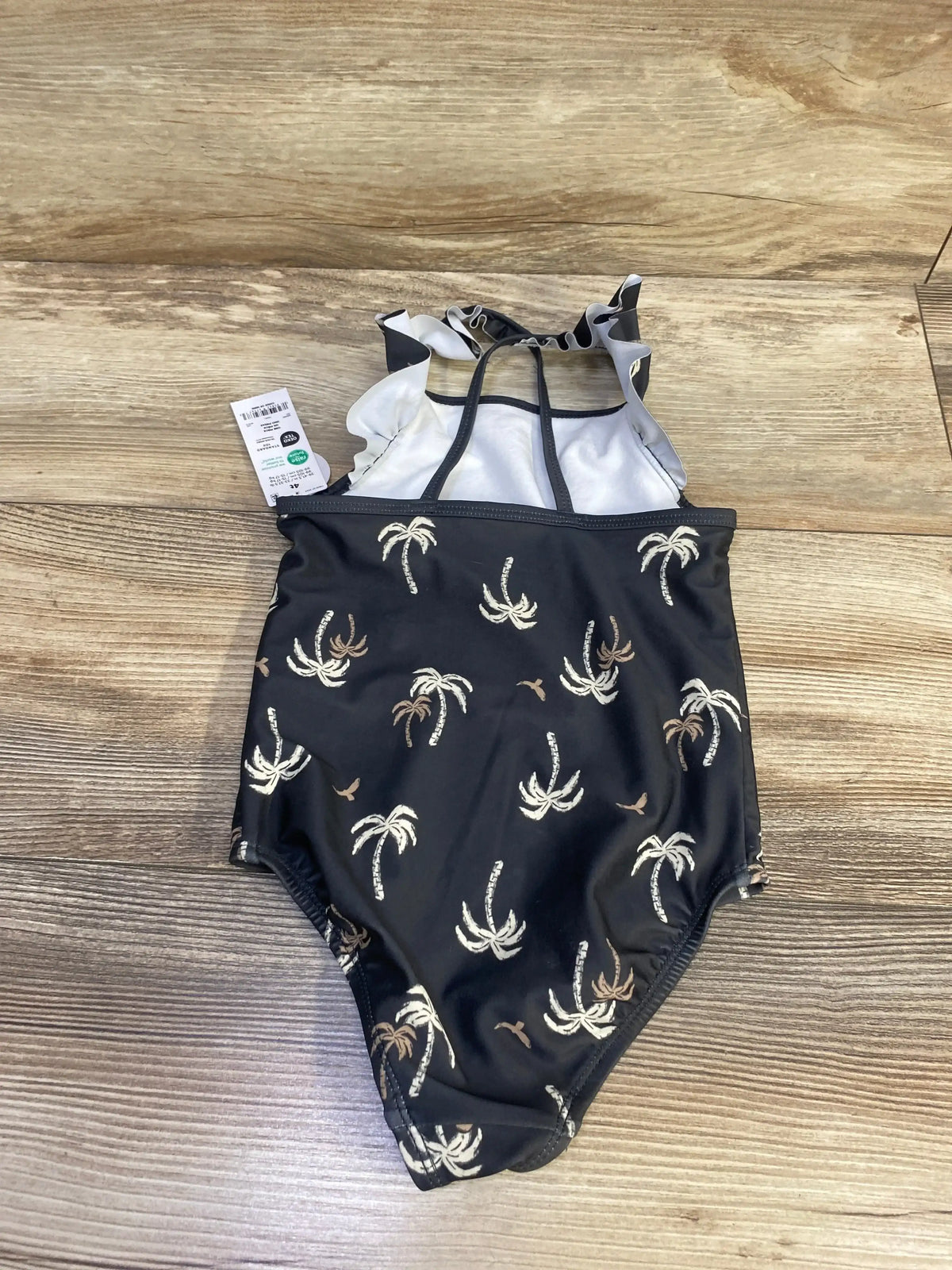 Front view of black Child Of Mine 1pc Palm Tree Swimsuit size 4T with palm tree print and ruffled straps on wood