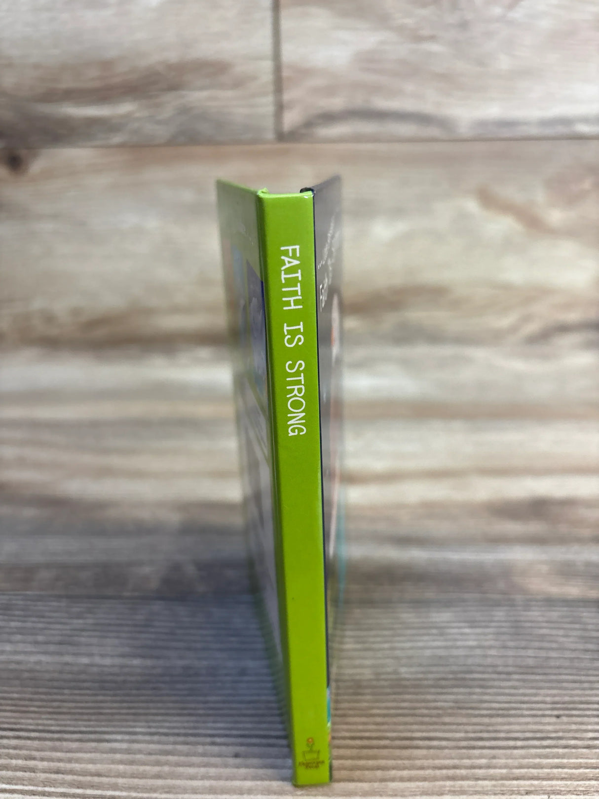 Spine view of Little Blessings Faith Is Strong Board Book in green standing upright on a wood plank background
