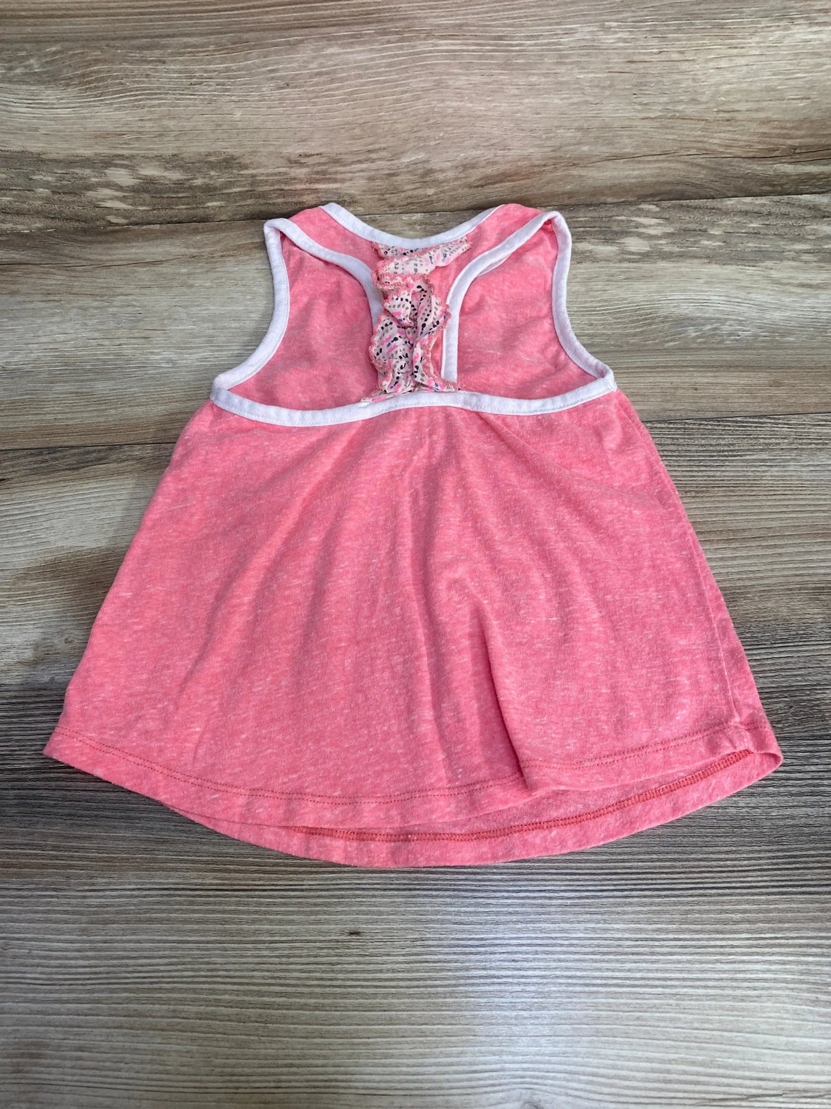Flat lay of Miki Miette pink toddler racerback tank top showcasing white trim and lace butterfly detail on the back on a wood surface