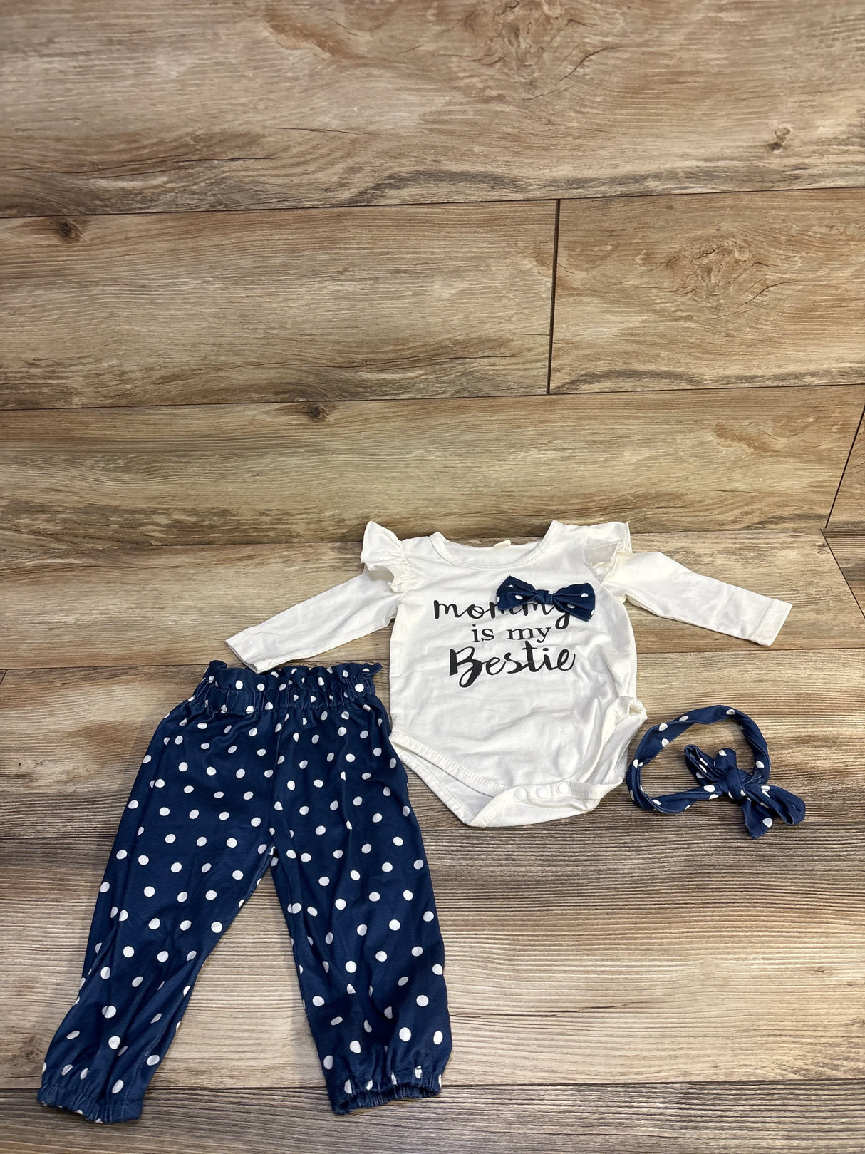 Flat lay of baby 3-piece outfit: white bodysuit reading Mommy is my Bestie, navy polka-dot pants and headband on wood