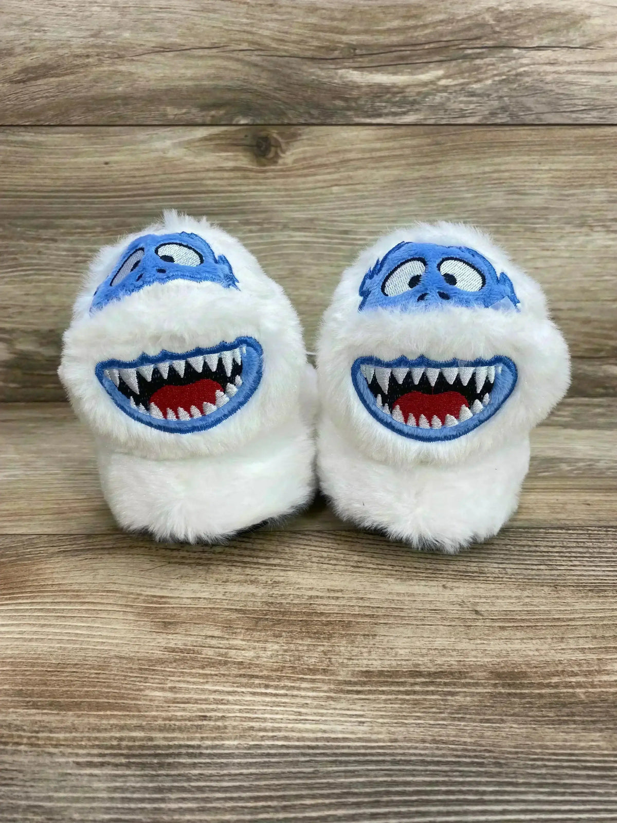 Front view of white plush Bumble the Abominable Snowman toddler slippers with embroidered blue face on wood background