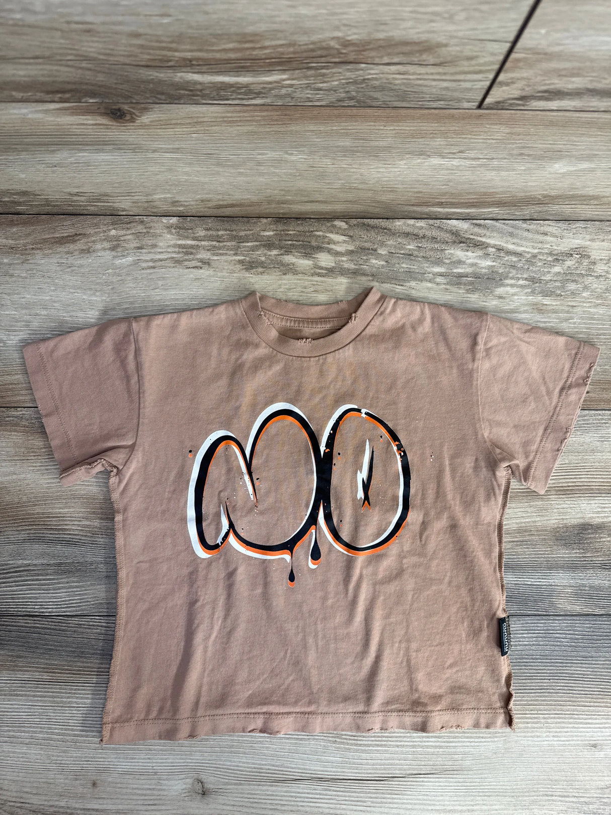 Brown kids t-shirt with black, white, and orange graffiti-style design on wooden floor