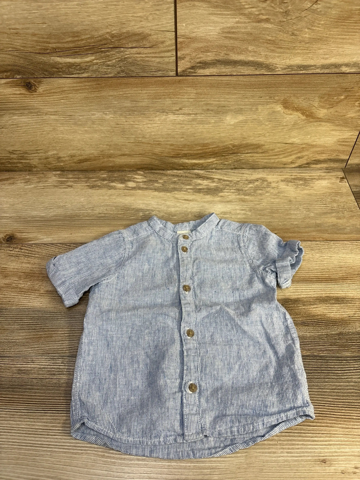 Flat lay of H&M blue and white striped short-sleeve button-up shirt showcasing front wooden buttons, size 12 months, on wood floor background