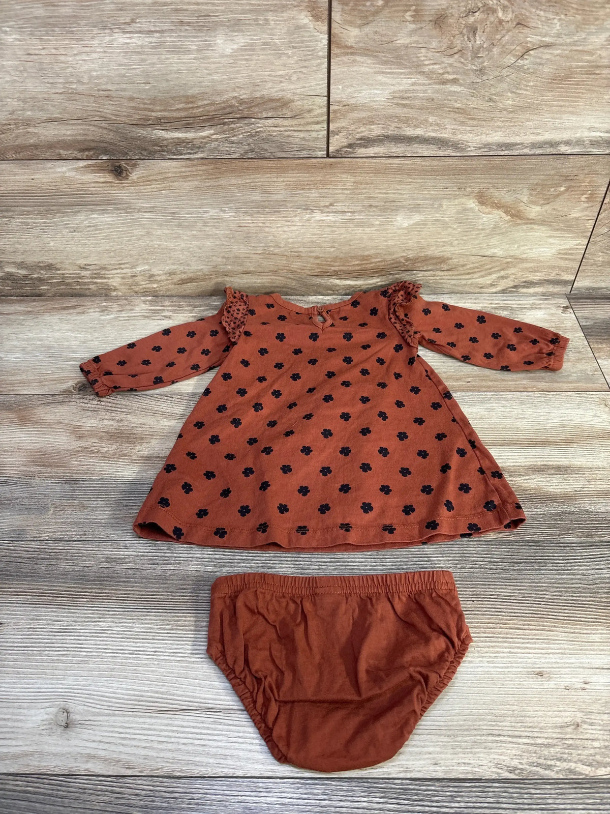Flat lay of brown baby dress with ruffled shoulders and black floral print, with matching bloomers on wood background