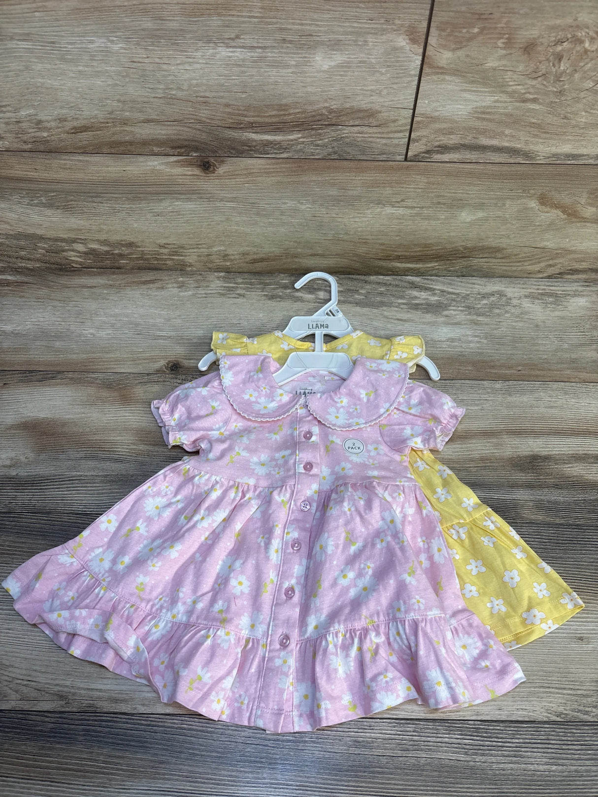 Two toddler girls' floral dresses, pink and yellow, short sleeve, displayed on wooden background