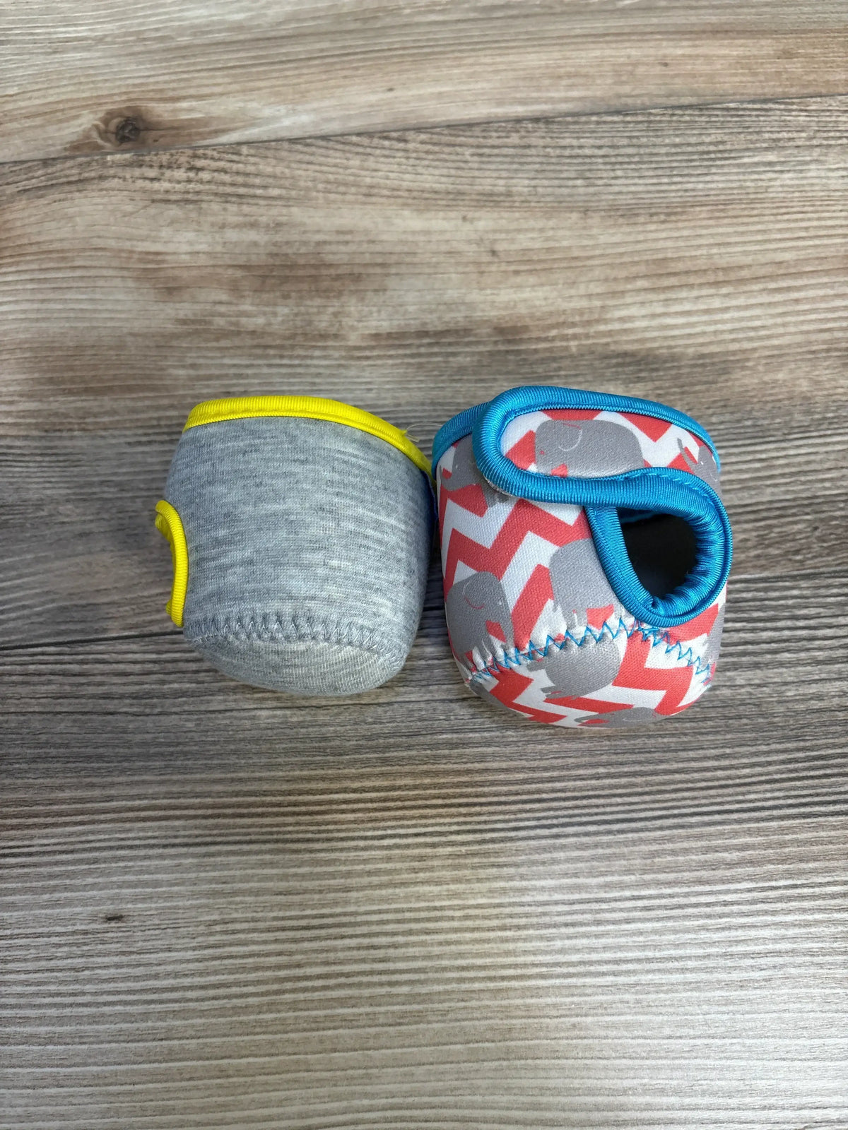 Top view of Miracle Bean Neoprene Baby Bottle Sleeves 2pc showcasing grey sleeve with yellow trim and red shark print sleeve with blue handle on wood plank backdrop