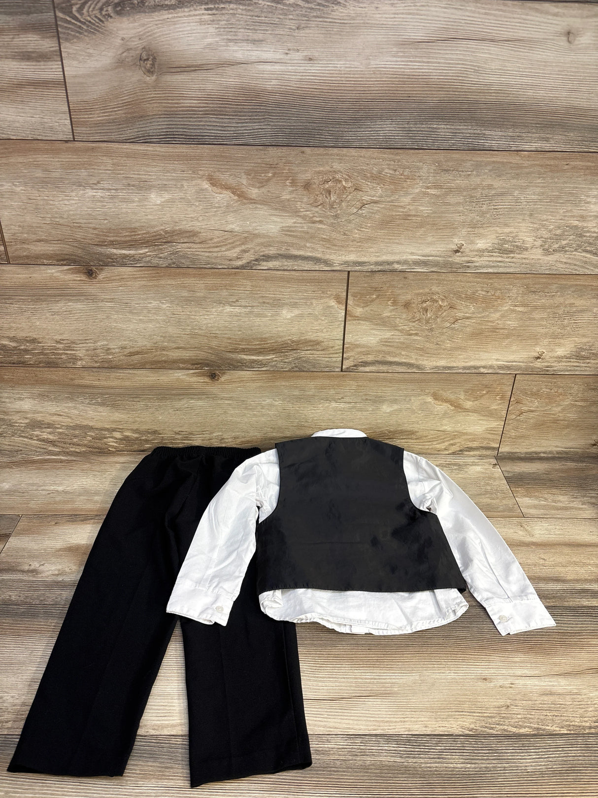 Kids formal outfit with black pants, white shirt, and black vest laid on wooden floor