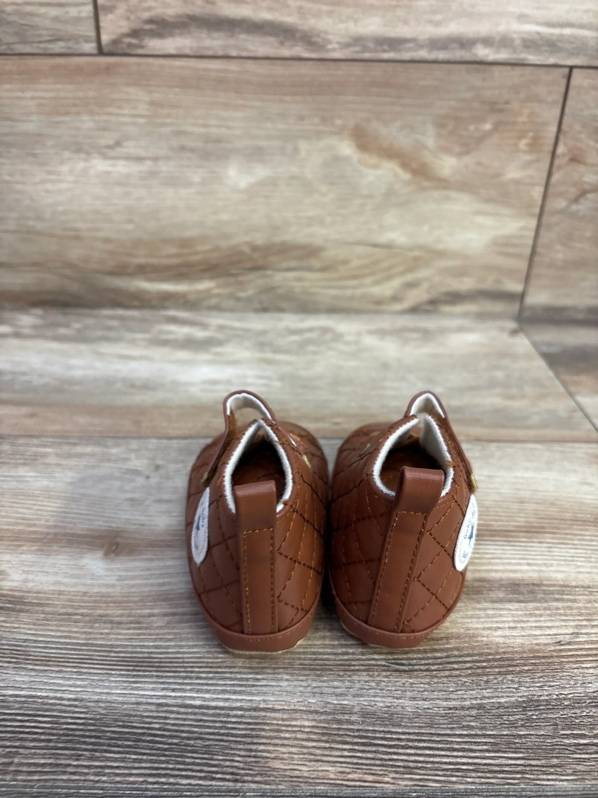 Rear view of brown faux leather crib shoes in size 4C showcasing diamond stitching and pull tabs on a wood-look floor