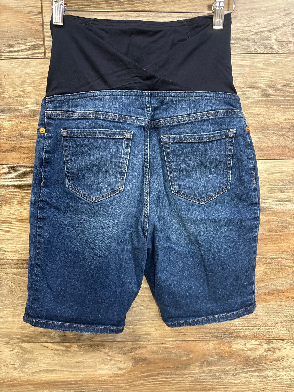 Back view of maternity denim Bermuda shorts with full black belly panel, back pockets, knee-length hem on wood background