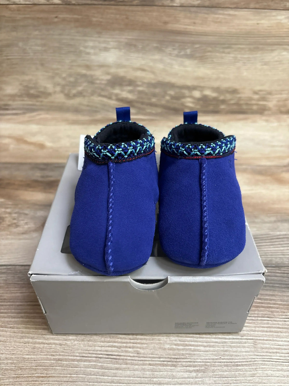 Front view of Ugg Baby Tasman blue suede slippers showing braided trim and pull tabs on a gray box over wood backdrop