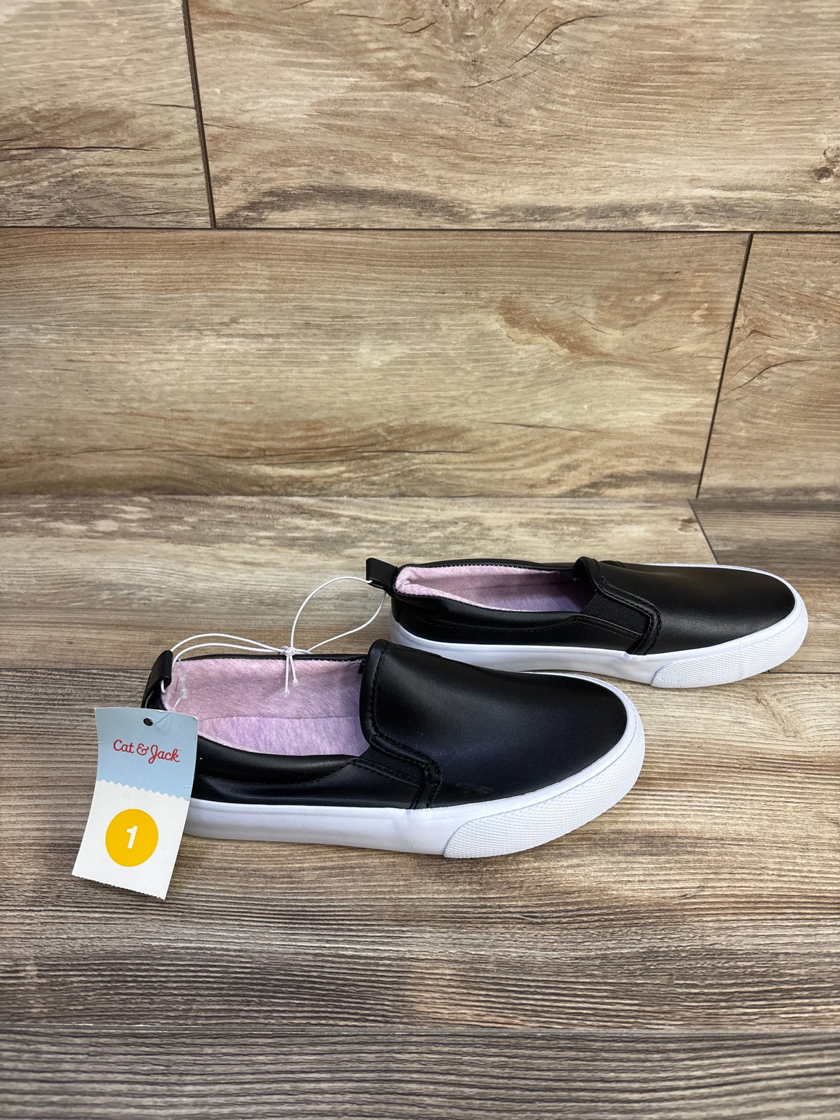 Side view of Cat & Jack Hailey toddler slip-on sneakers in black showing pink interior lining and white rubber sole on wood floor, size 1 tag visible