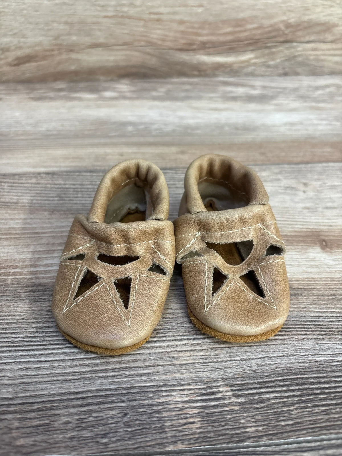 Top view of baby tan soft sole leather moccasins showcasing geometric cutout vamp and contrast stitching on wood plank floor
