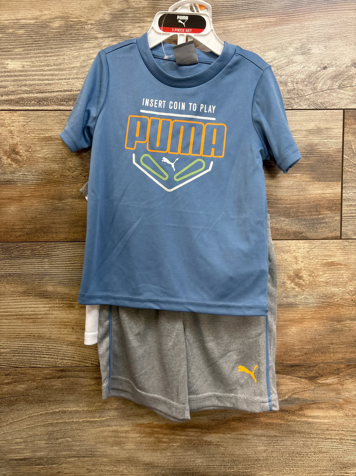 Front view of 3-piece Puma set: blue tee showing INSERT COIN TO PLAY, white tank, gray shorts with yellow Puma logo on wood