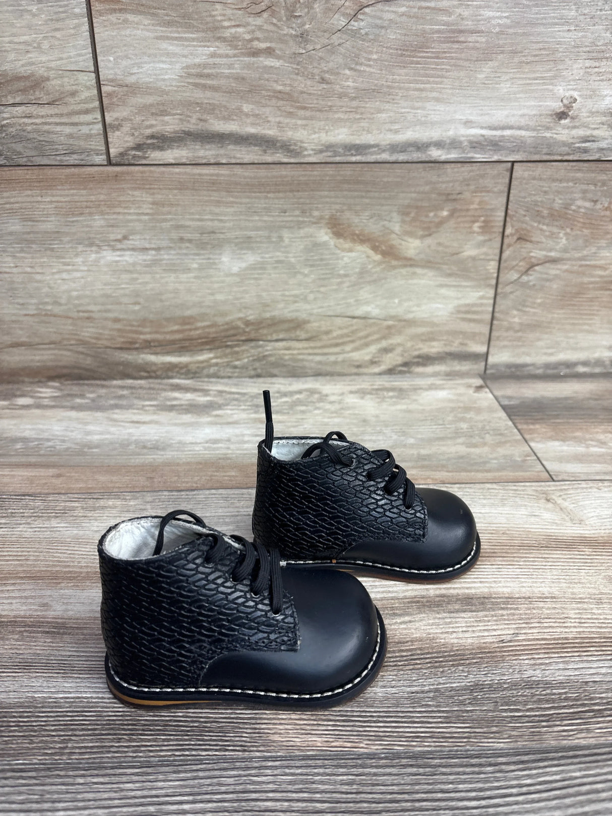 Three-quarter view of black Josmo First Walker Oxford shoes showcasing textured leather uppers and four-hole laces on a wood-look floor
