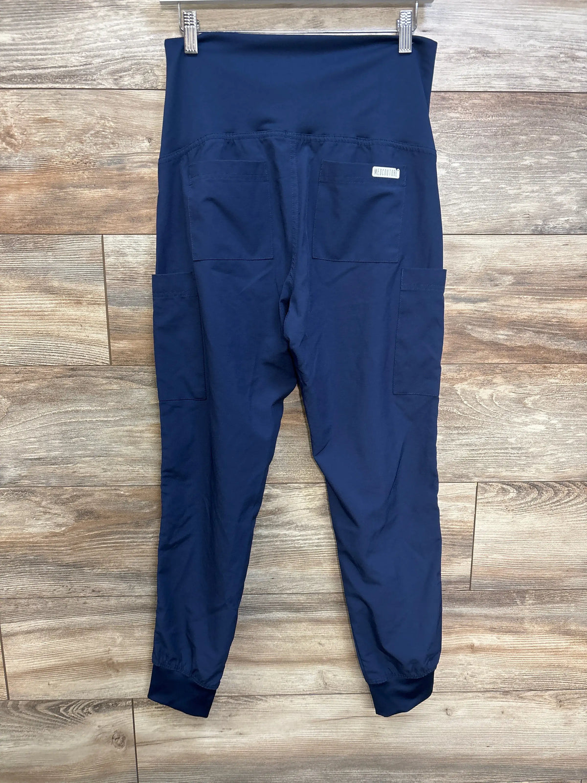 Flat lay of navy Medcouture maternity scrub pants in XS showcasing the full belly panel and multiple pockets on wood floor