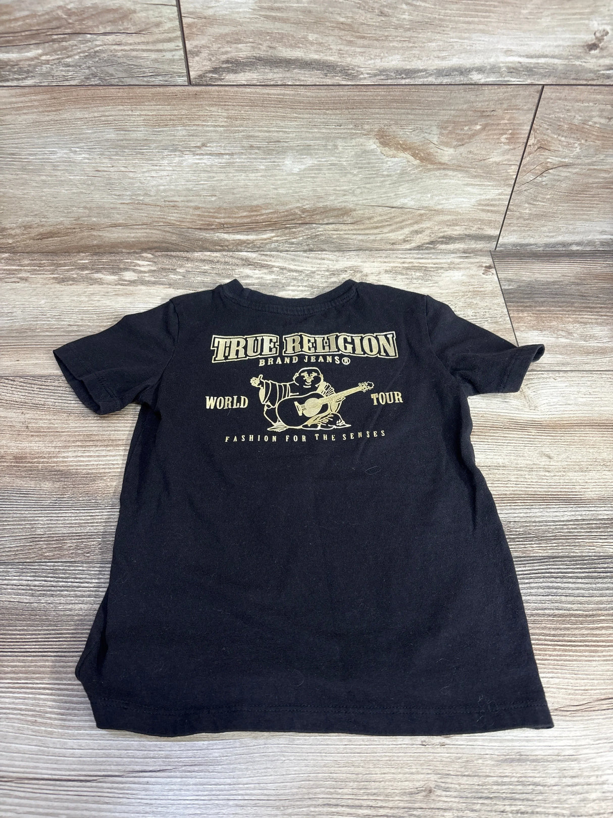 Black kids' True Religion graphic t-shirt with gold lettering, laid flat on wood floor.