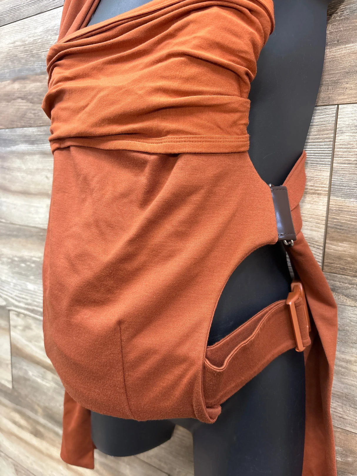 Rust orange fabric baby hip carrier with adjustable buckle on black mannequin against wood background