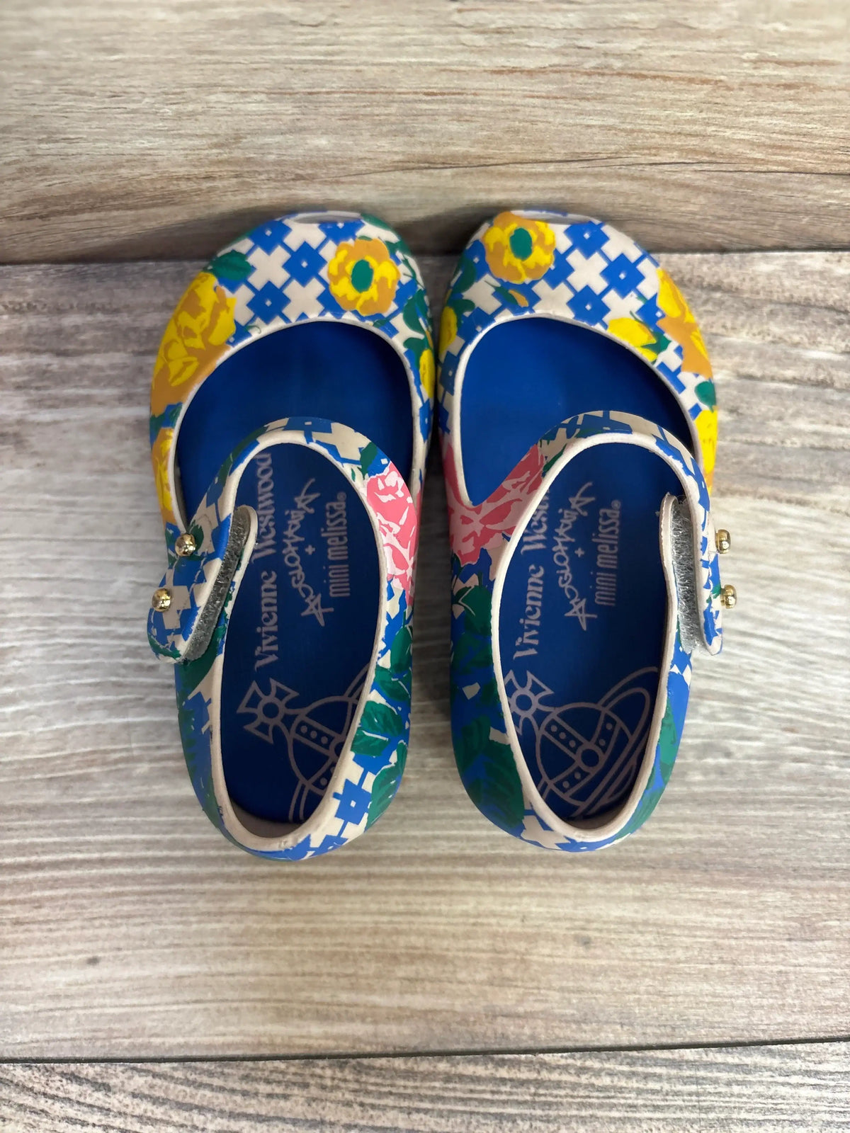 Top view of toddler Mary Jane shoes, blue with yellow floral and white geometric print, strap with gold studs on wood