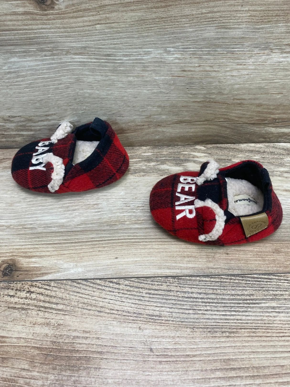 Angled view of Dearfoams baby slippers (size 4C) showing red and black plaid, BEAR embroidery and sherpa lining on wood