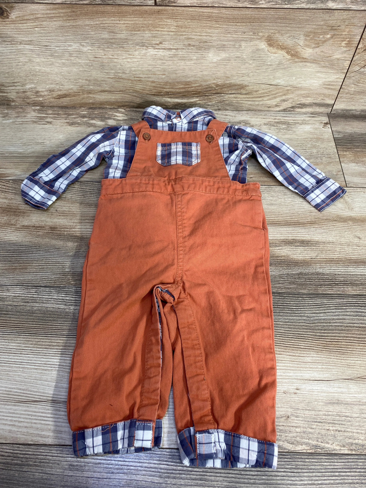Children's outfit with orange overalls and blue plaid shirt, laid flat on wood floor