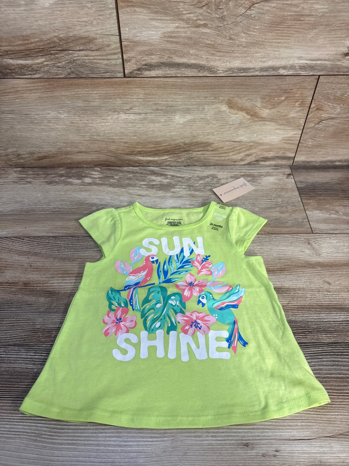 Front view of First Impressions lime Sun Shine shirt for 24 months with short sleeves and floral SUN SHINE graphic on wood floor