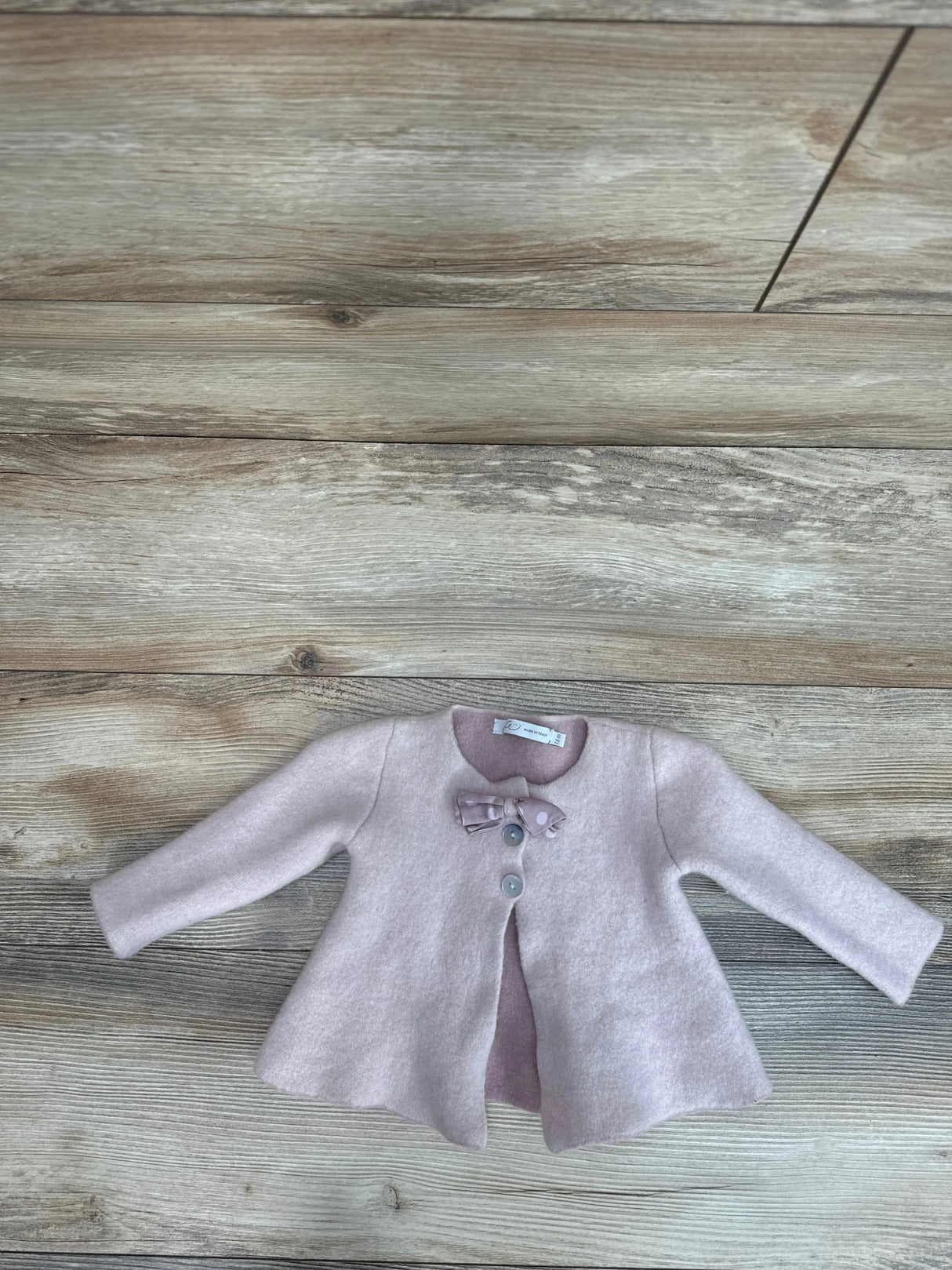 Flat lay of pink Per Te baby girl cardigan size 18 months with bow at neckline, two buttons and flared hem on wood floor
