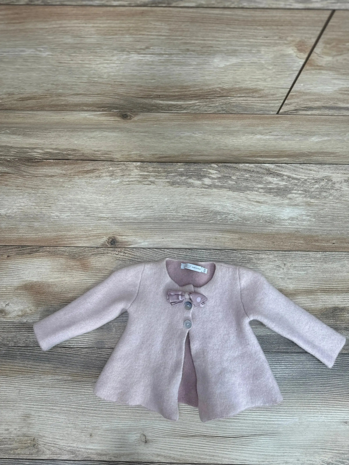 Flat lay of pink Per Te baby girl cardigan size 18 months with bow at neckline, two buttons and flared hem on wood floor