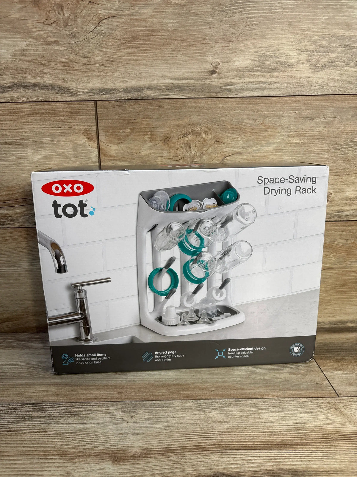 Front view of NEW OXO TOT Space Saving Drying Rack packaging showcasing angled pegs for bottles and cups with a wood floor background