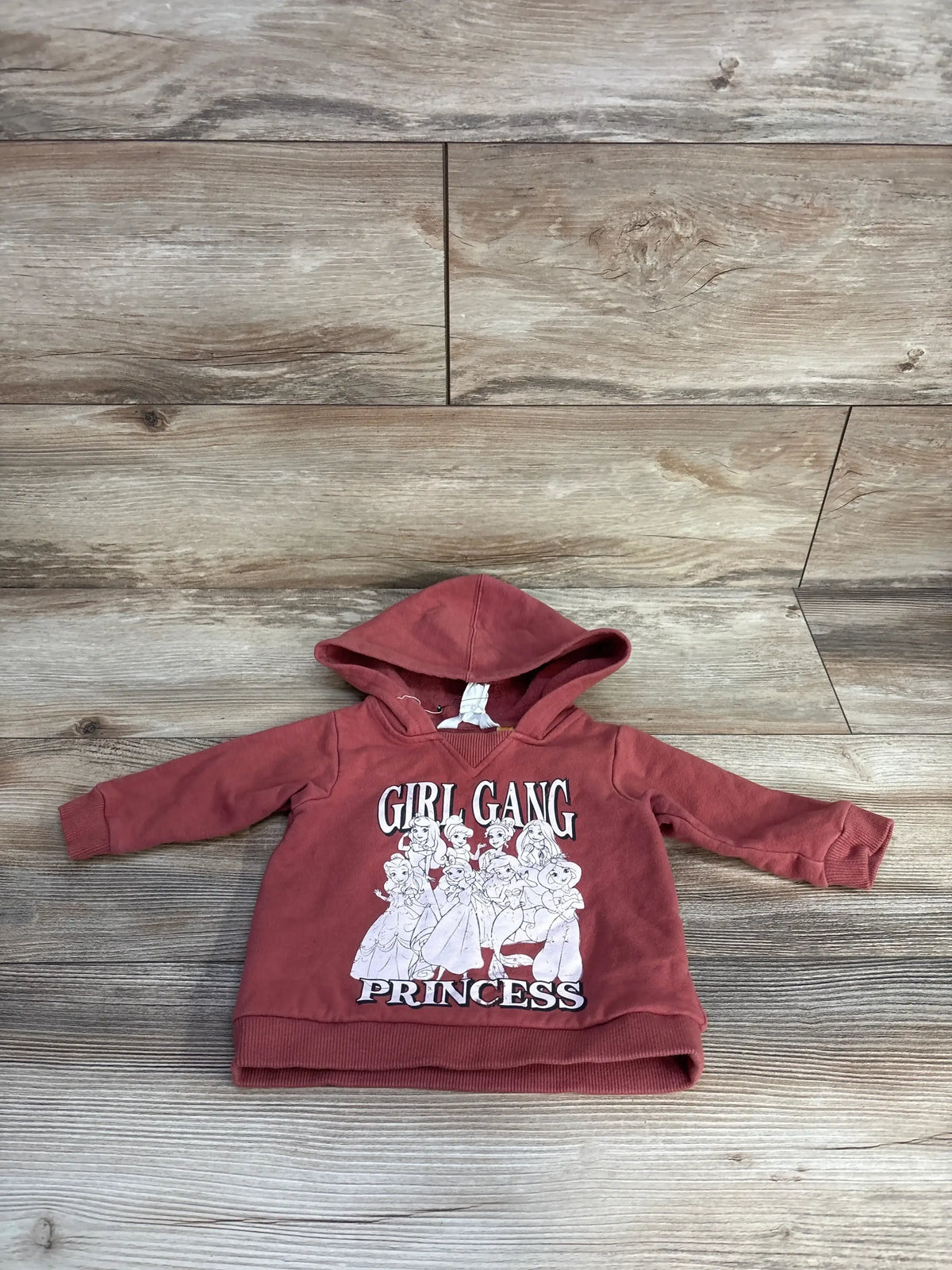 Flat lay of red toddler Cotton-On pullover hoodie showing Disney princess graphic with ‘GIRL GANG’ and ‘PRINCESS’ text on wood floor