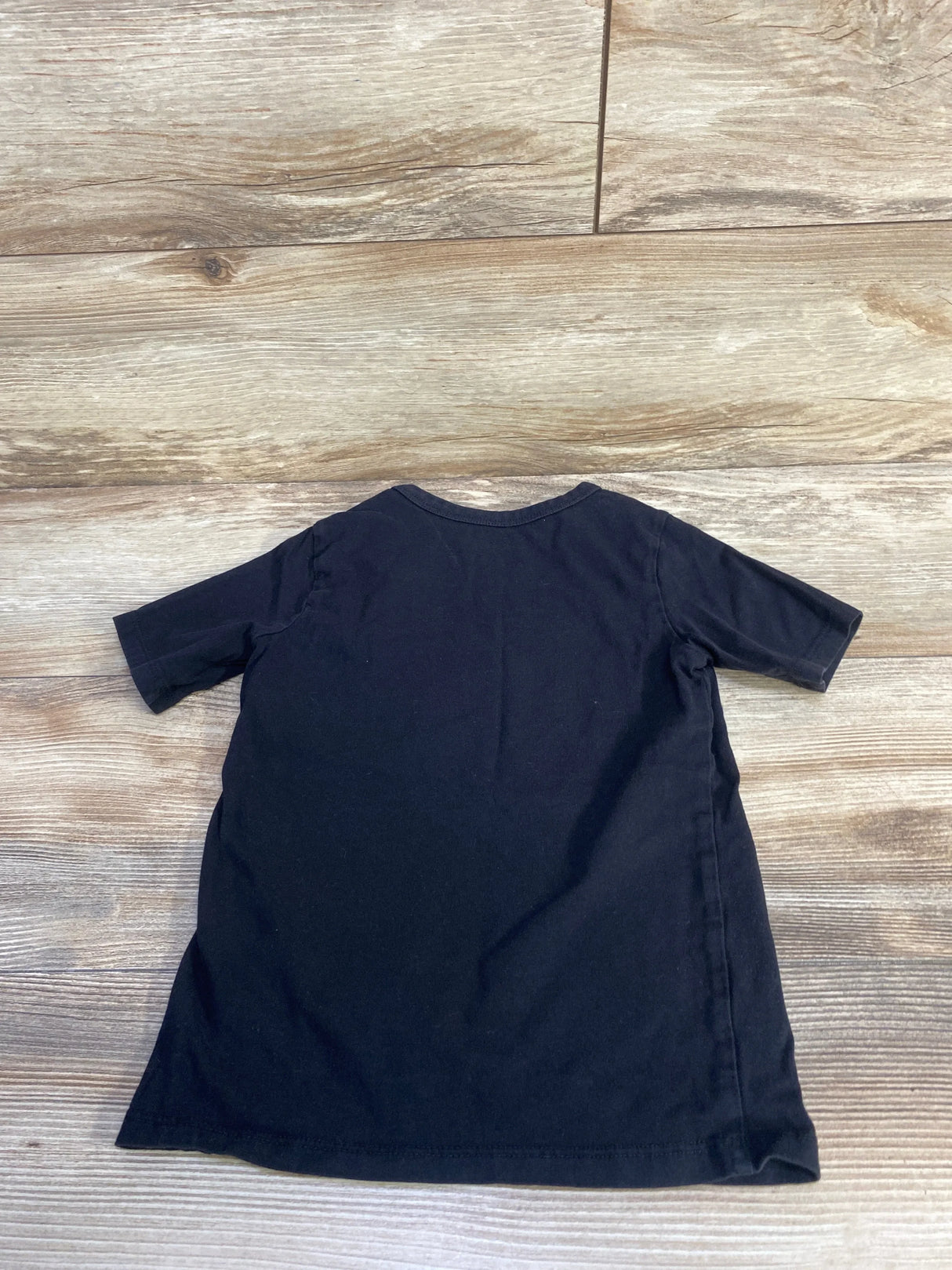 Back view of children’s black short-sleeve t-shirt showcasing crew neckline and plain fabric on light wood floor