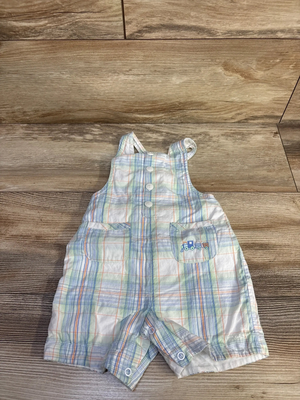 Front view of white pastel plaid shortalls with three buttons, pockets with train embroidery and snap crotch on wood floor