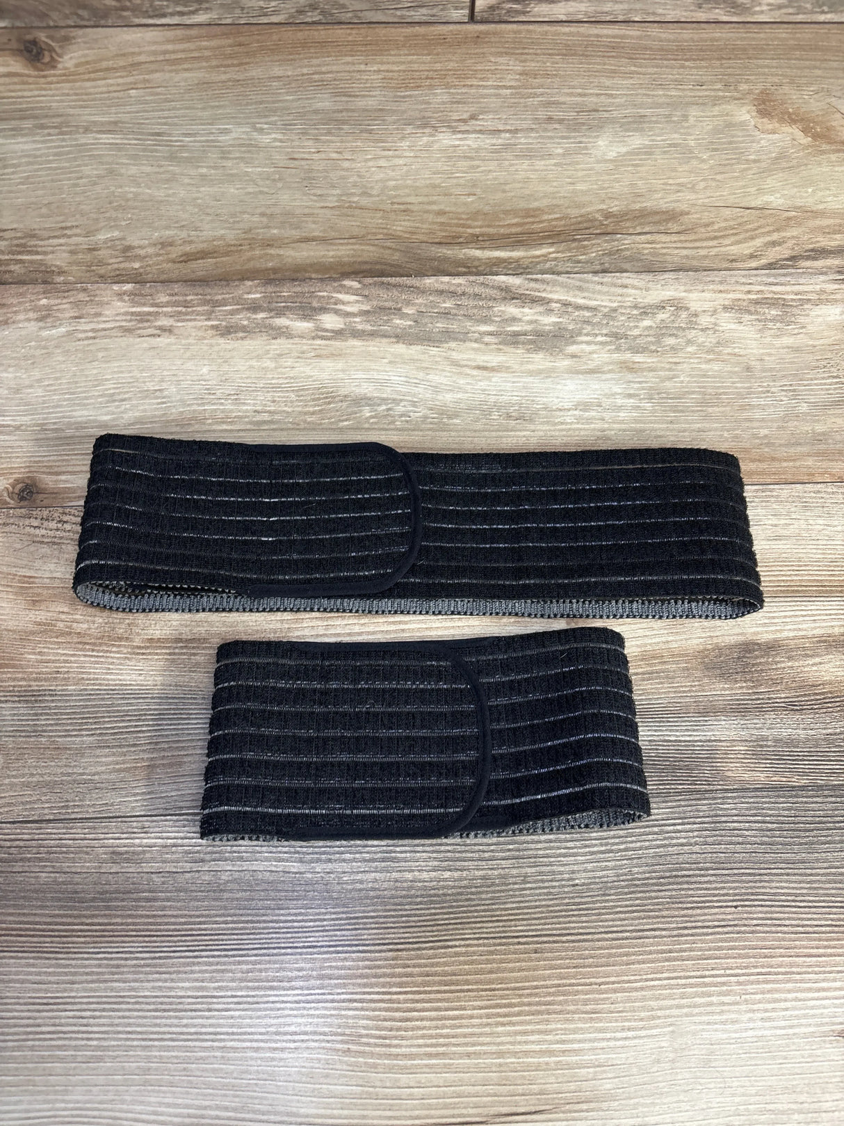 Top-down view of Belly Bandit 2-in-1 Hip Bandit black showing two ribbed elastic bands (hip and belly) with Velcro on wood