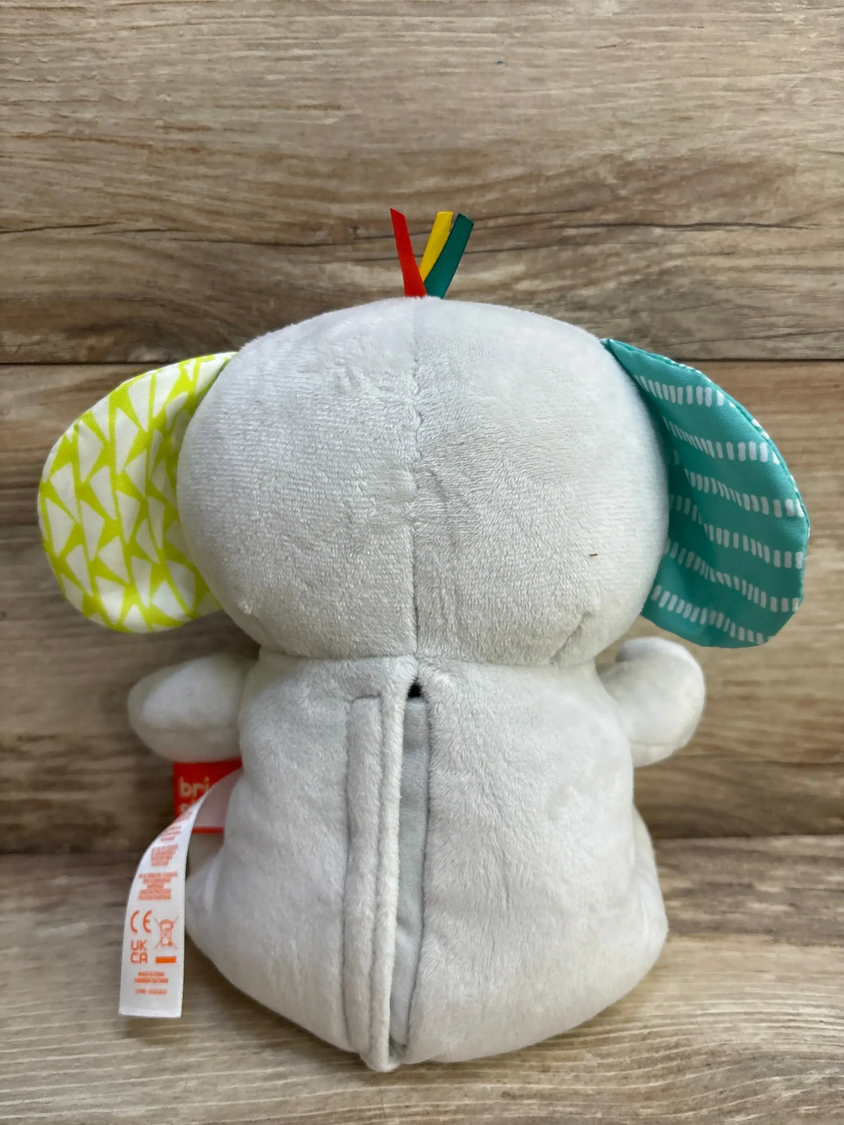 Back view of Bright Starts Hug-a-Bye Baby Elephant plush with yellow and teal ears, ribbon tags, and rear seam on wood