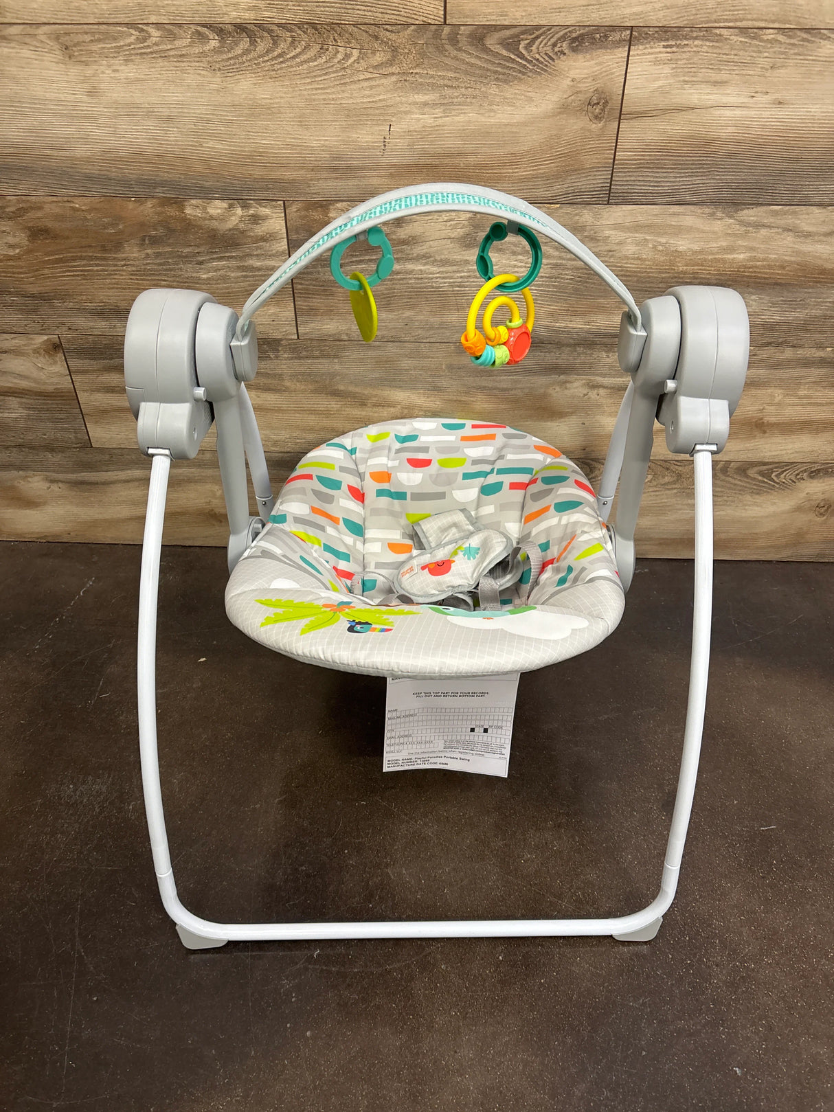 Front of Bright Starts portable compact baby swing with patterned seat, toy bar with two toys, harness, gray frame, wood wall