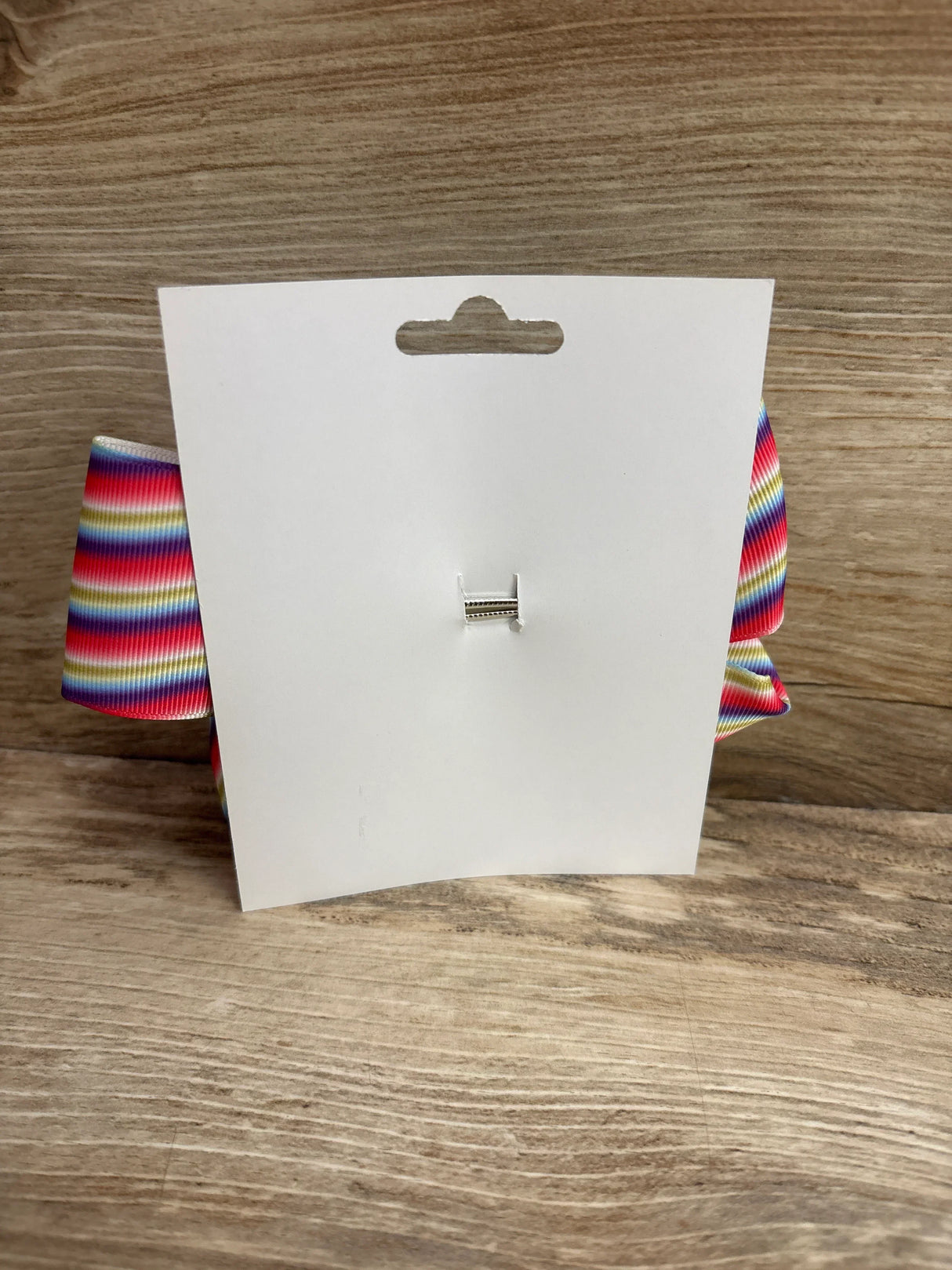 Back view of large striped hair bow showing horizontal rainbow stripes and metal clip on a white display card against wood background