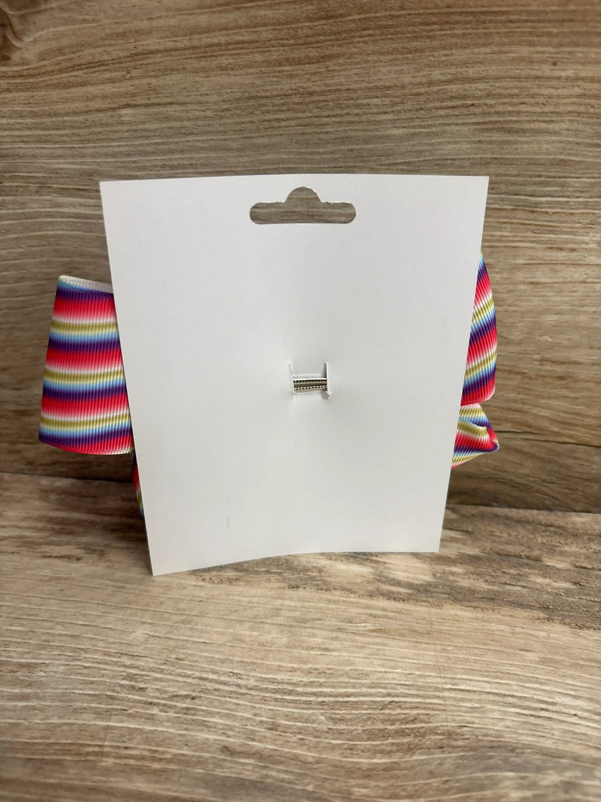 Back view of large striped hair bow showing horizontal rainbow stripes and metal clip on a white display card against wood background