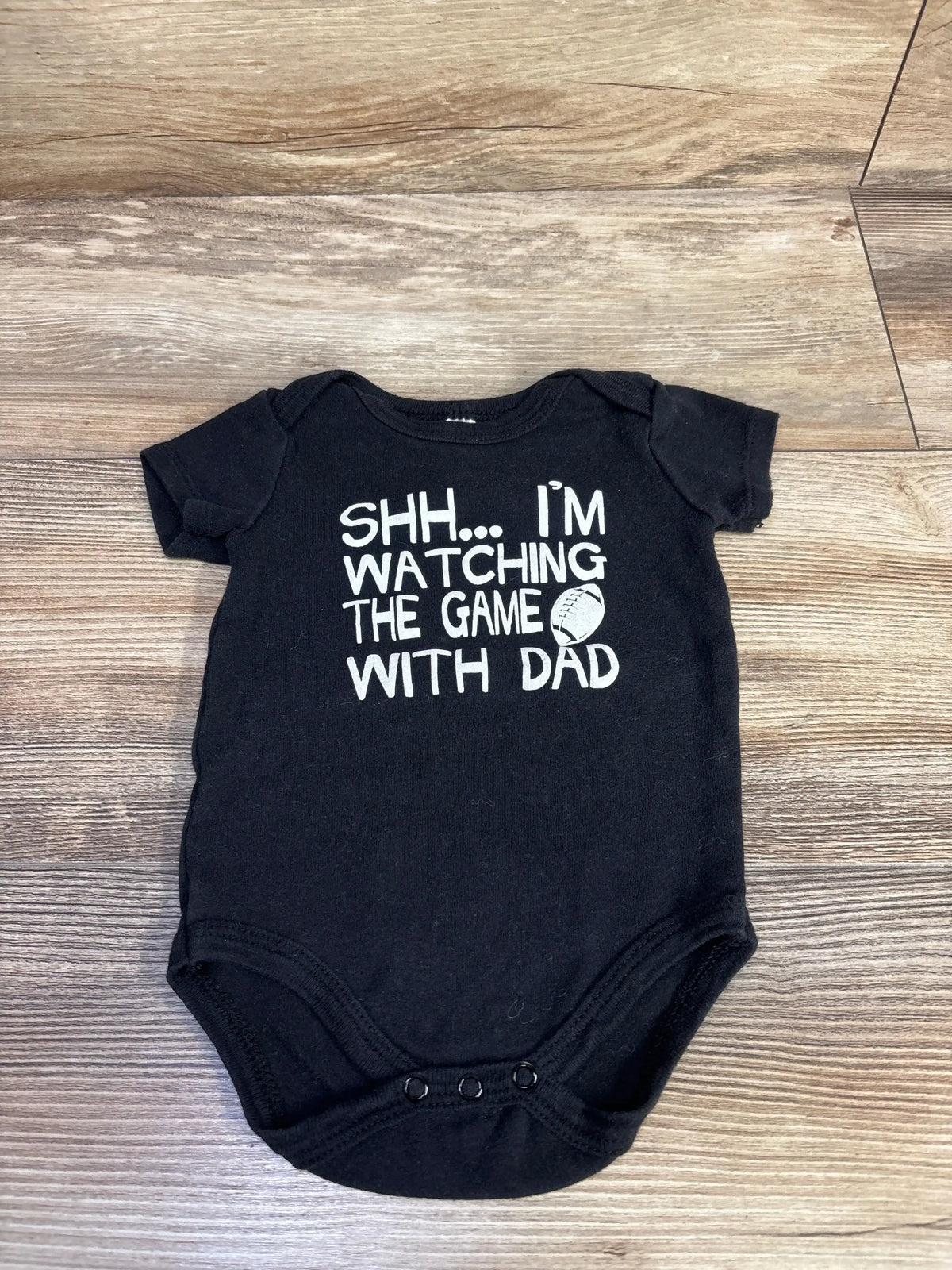 Flat lay of black bodysuit with white text ‘Shh... I’m watching the game with Dad’ and small football icon on wood background