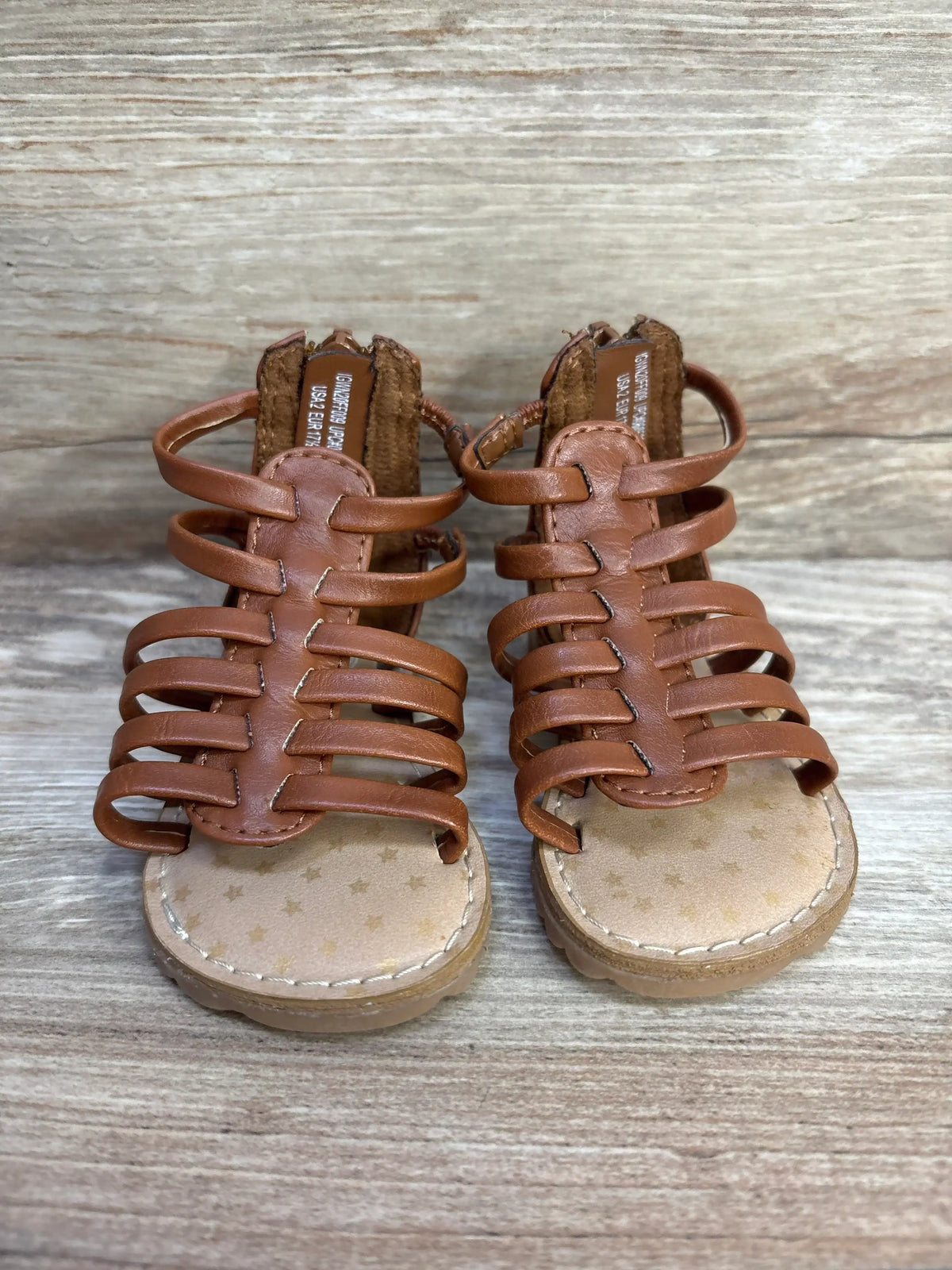Front view of brown toddler gladiator sandals showing multiple straps, back zipper, and star-print insole on wood background