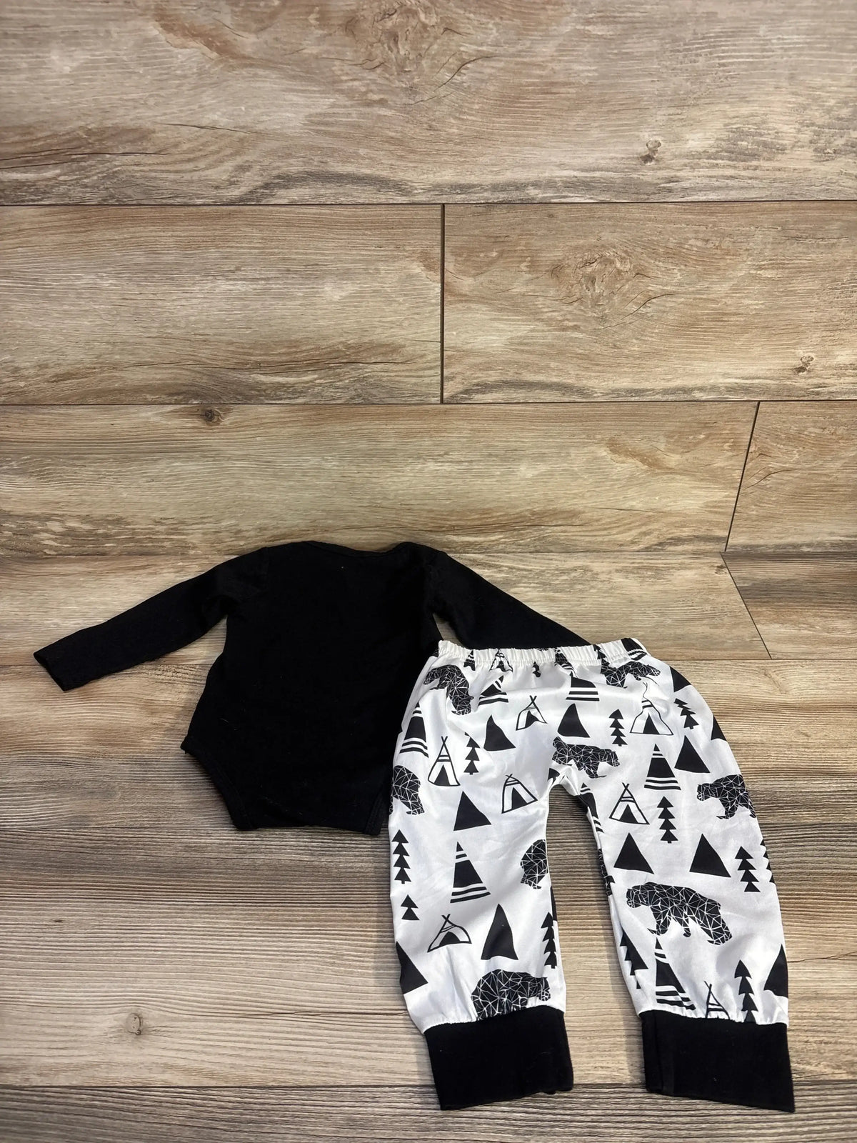 Flat lay of black long-sleeve bodysuit and white elastic-waist pants with black teepee and bear print on wood plank background