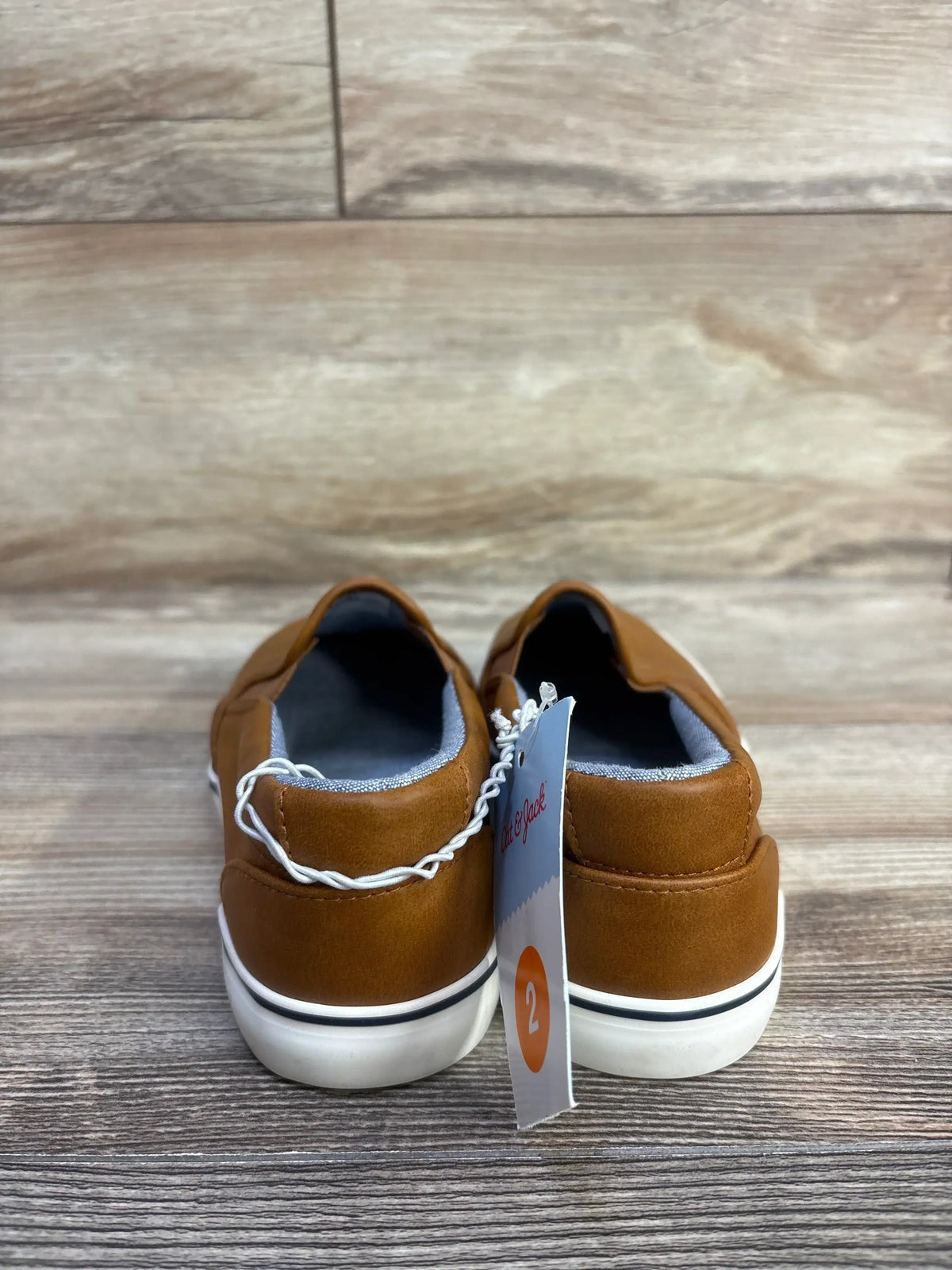Rear view of Cat & Jack Enzo toddler slip-on sneakers in tan showing white rubber soles with black stripe and size 2 tag on wood floor